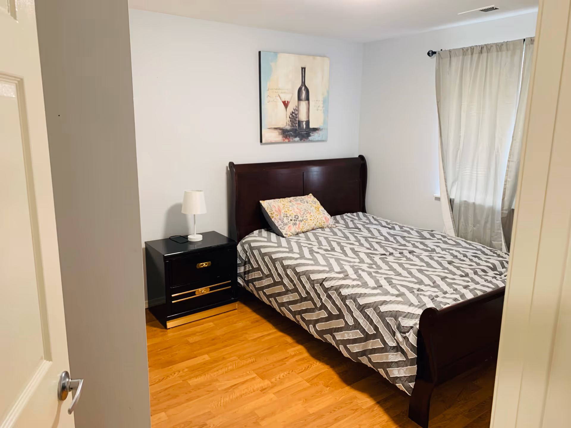 A bedroom with a wooden bed frame and a gray and white patterned bedspread. There is a floral pillow on the bed. Next to the bed is a black nightstand with two drawers and a white lamp on top. A painting of a wine bottle and glass hangs on the wall above the bed. A window with beige curtains is on the right side of the room. The floor is wooden.