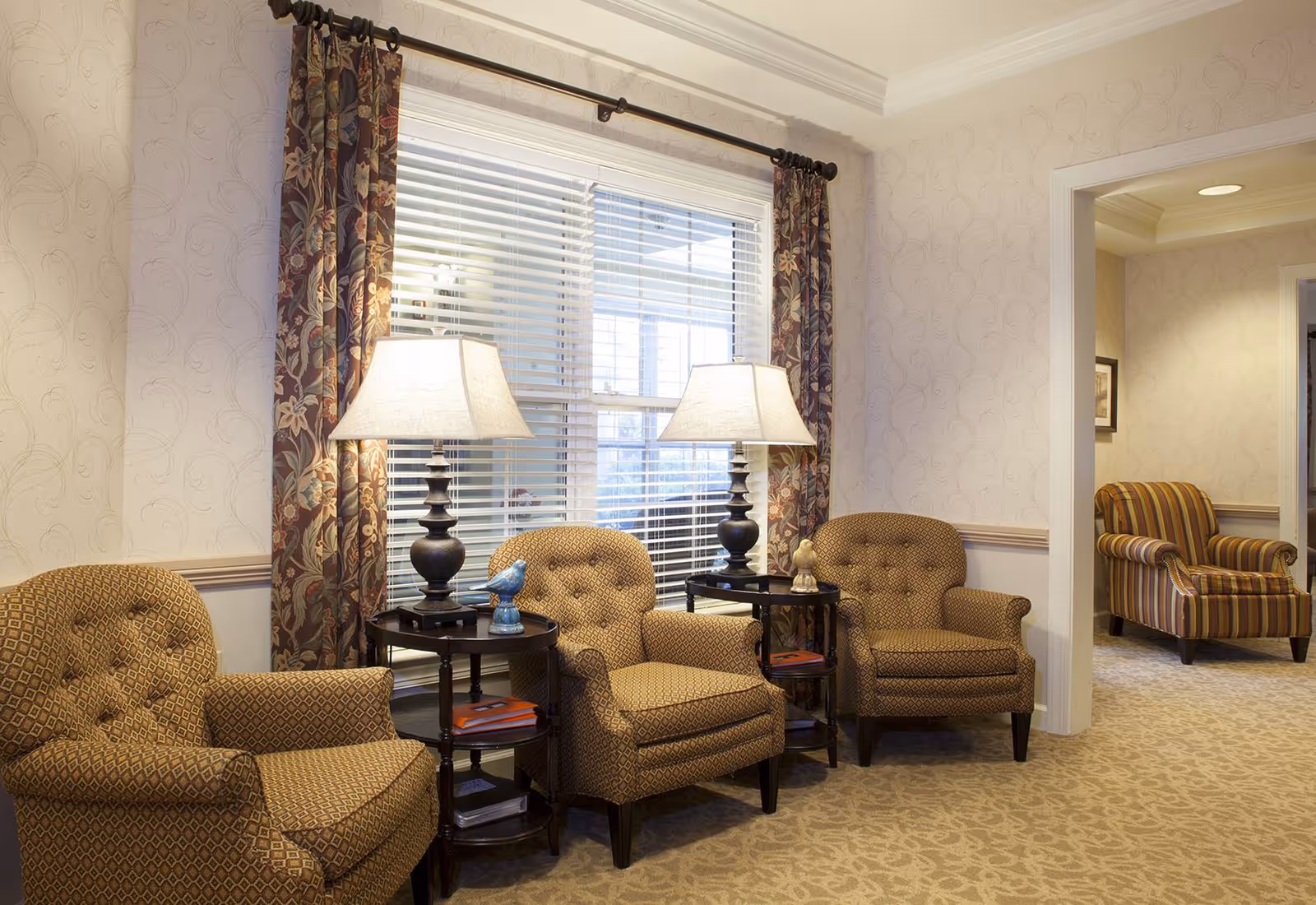 A cozy sitting area with three patterned armchairs arranged around two small round tables, each holding a lamp and decorative figurines. The area is well-lit by natural light coming through a window with floral curtains and white blinds. In the background, another striped armchair is visible in an adjacent room with similar wallpaper and carpet.