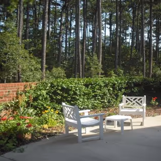 Two white outdoor chairs and a small table on a patio next to a garden and low brick wall with tall pine trees beyond.