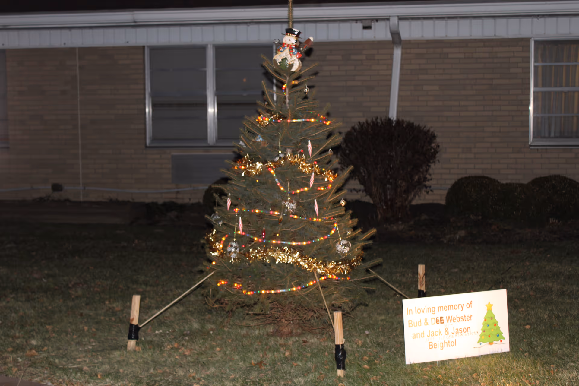 A small decorated Christmas tree outdoors at night with lights, candy cane ornaments, gold garland, and a snowman tree topper. The tree is supported by wooden stakes and black tape. Behind the tree is a brick building with windows. A sign in front of the tree reads, 'In loving memory of Bud & Dee Webster and Jack & Jason Beightol' with a small Christmas tree graphic.