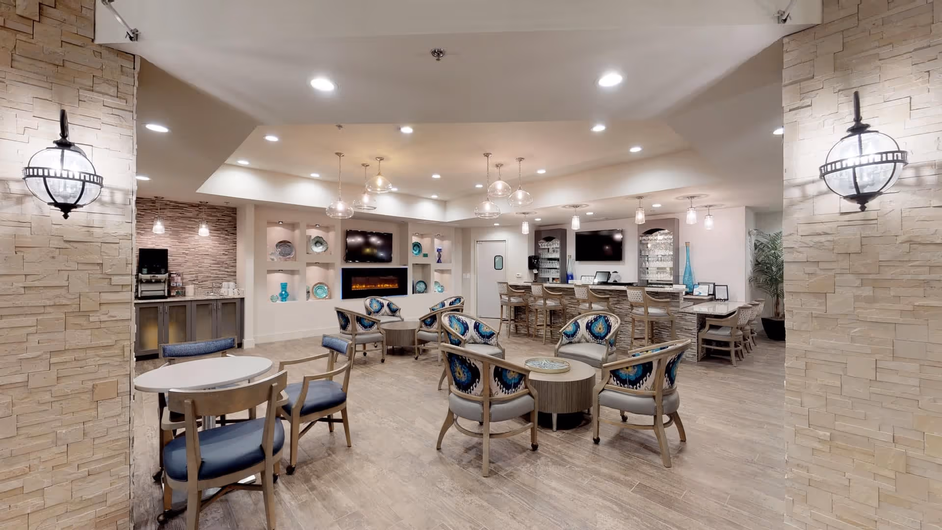 A spacious and well-lit common area in a senior living facility featuring multiple seating arrangements with chairs and small tables. The room has modern decor with stone accent walls, pendant lighting, and built-in shelves displaying decorative plates and vases. There is a bar area with stools and a coffee station in the background.