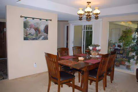 A dining room with a wooden table and six matching chairs. The table is set with placemats and a floral centerpiece. A chandelier with multiple lights hangs above the table. There is a wall hanging with a scenic image on the left wall and a large window or glass door on the right side showing some greenery outside.