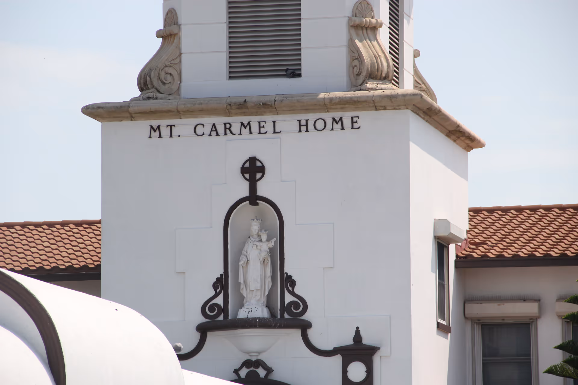 Close-up view of the exterior of a building with a white facade and red tile roof. The building has a decorative statue of a religious figure in a niche, with the words 'MT. CARMEL HOME' displayed above it.