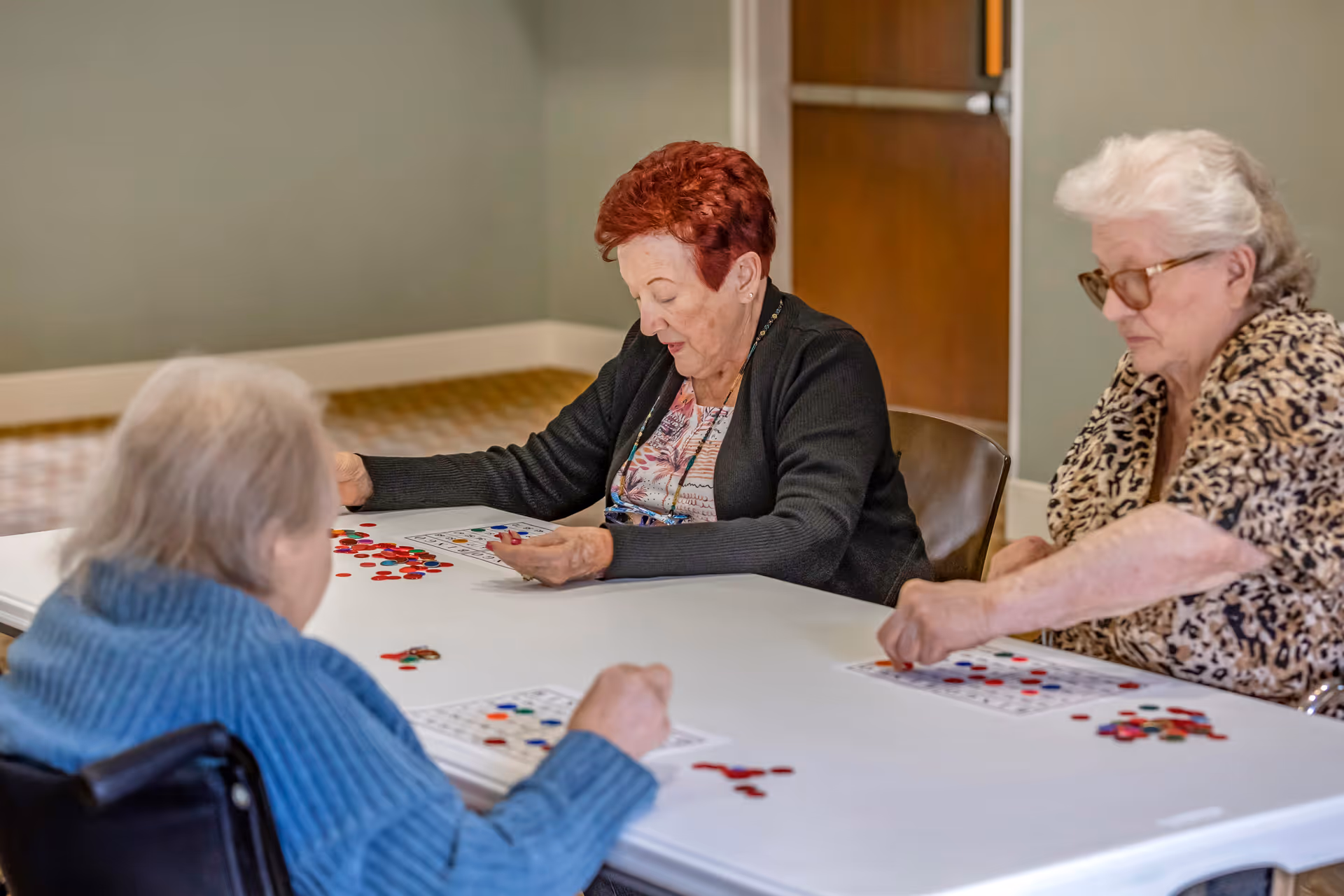 Three elderly women sitting around a white table playing a game with colorful chips on bingo cards in a room with light green walls and a brown refrigerator in the background.