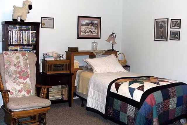 A cozy bedroom with a single bed covered with a white quilt and a colorful patchwork blanket. Next to the bed is a wooden nightstand with a lamp and decorative items. A wooden rocking chair with a floral cushion is placed near a bookshelf filled with DVDs and books. The walls are decorated with framed pictures and certificates.