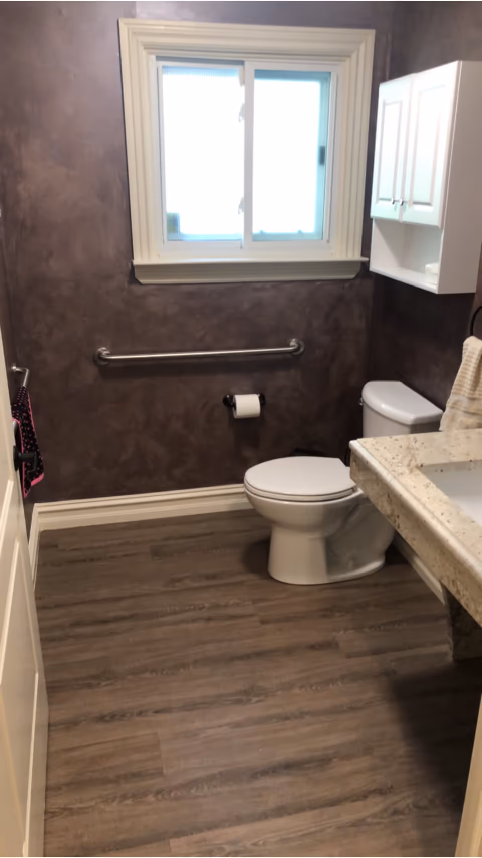 A bathroom with a white toilet, a granite countertop with a sink, a white wall cabinet, a window with frosted glass, a grab bar on the wall, and wood-look flooring.