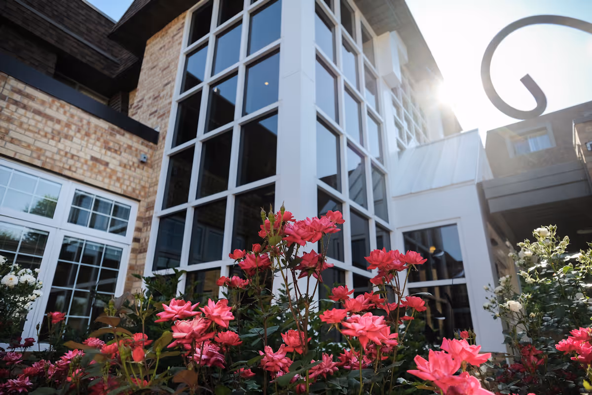 Exterior view of a building with large windows and brick walls, surrounded by blooming pink and white flowers in the foreground, with sunlight shining from the top right corner.