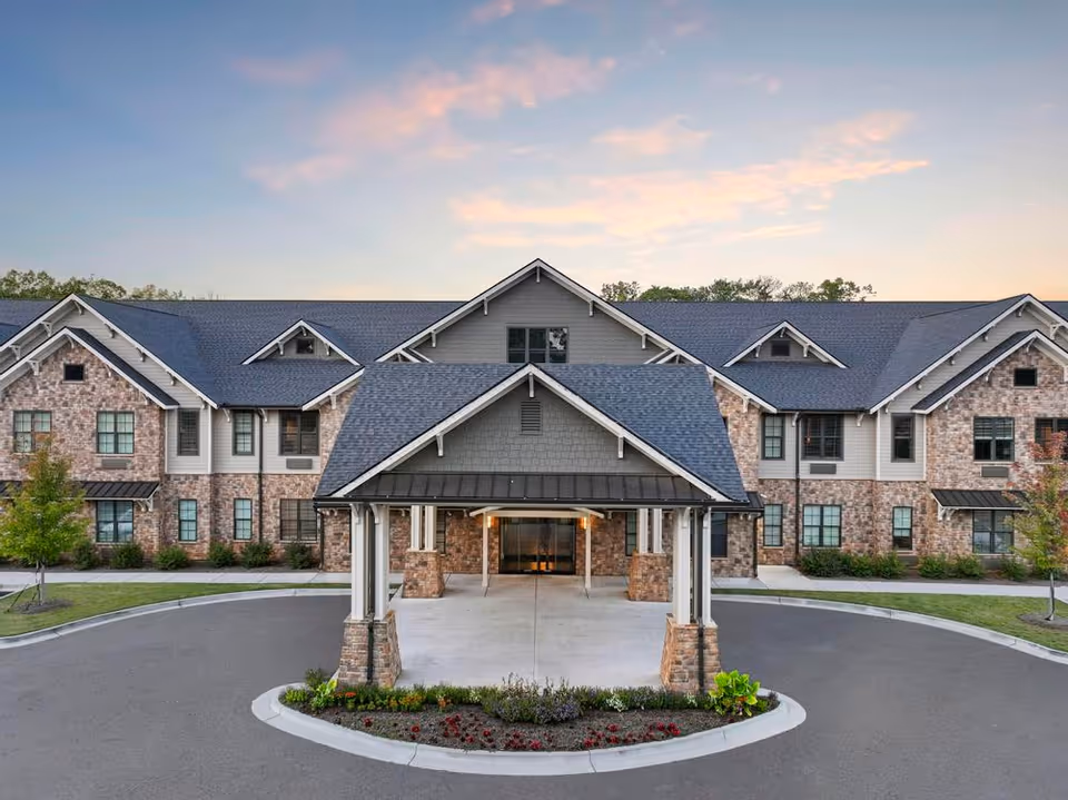 Front entrance with a covered porte-cochère of a two-story brick senior living building under a pastel sky.