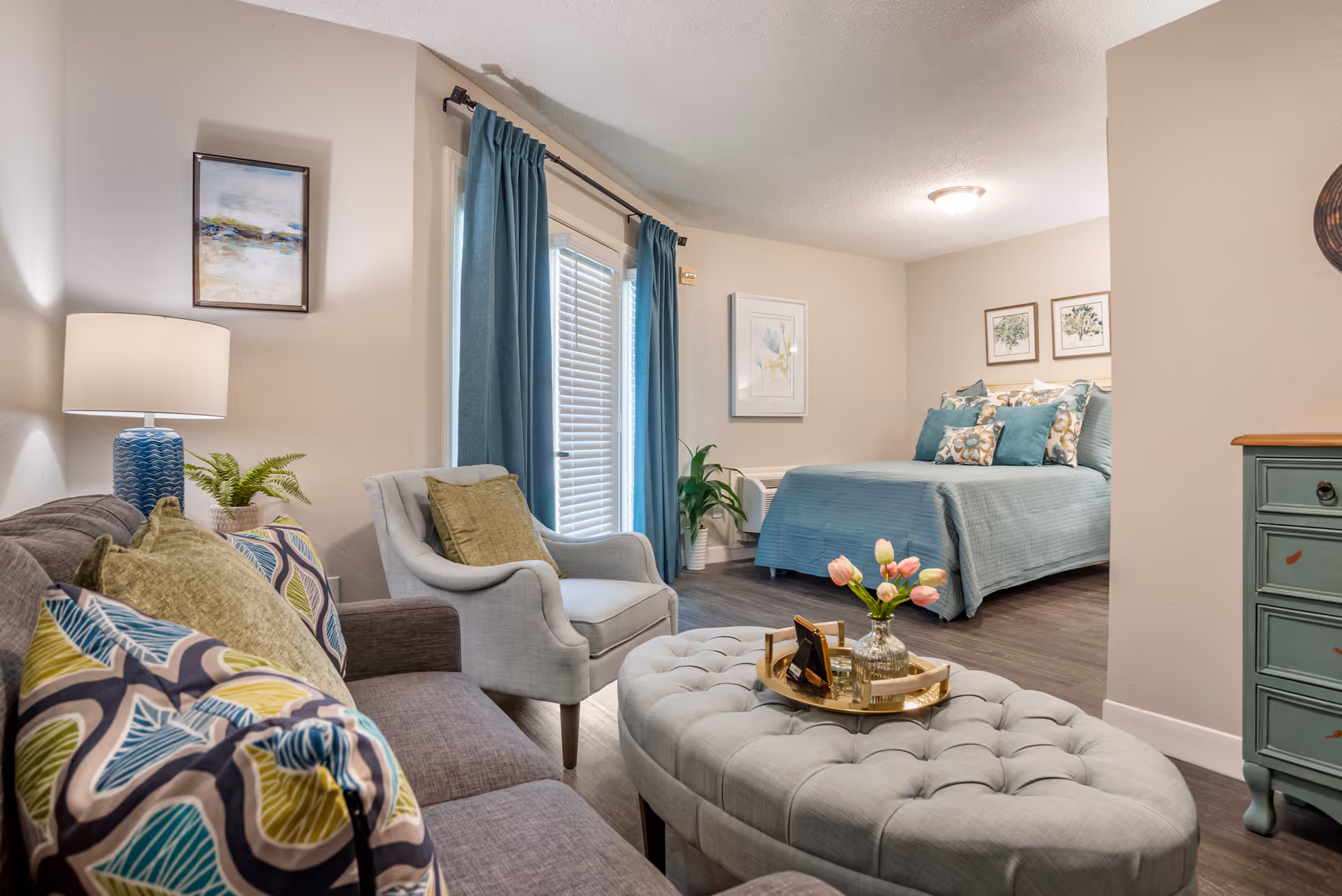 A cozy living and bedroom area in a senior living facility. The living room features a gray sofa with colorful patterned pillows, a light gray armchair, a tufted ottoman with a tray holding a vase of pink tulips, and a blue table lamp on a side table. The bedroom area has a bed with a blue bedspread and multiple pillows, framed artwork on the walls, and blue curtains covering a window or door.
