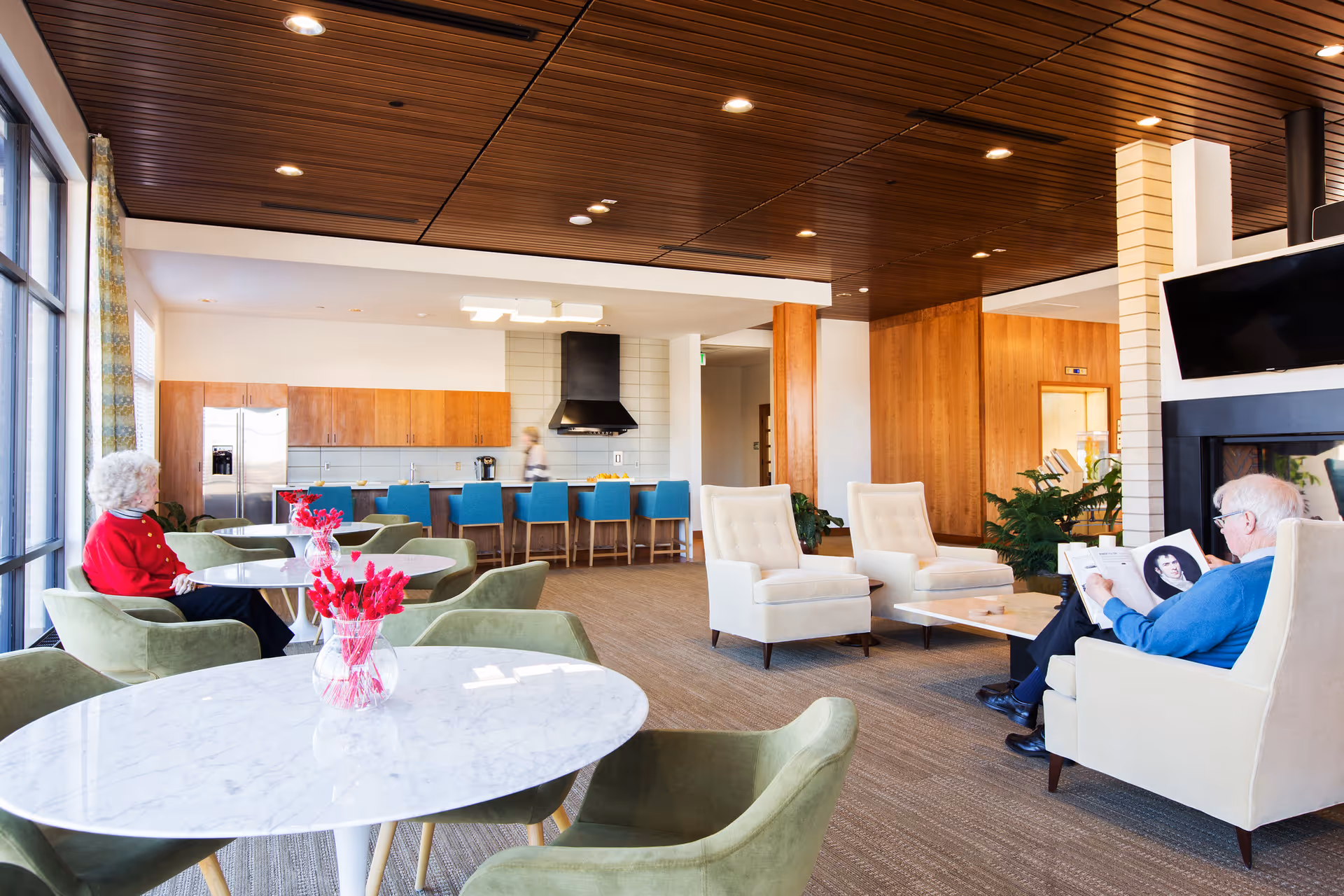 A spacious senior living common area with modern decor featuring a kitchen with wooden cabinets, a stainless steel refrigerator, and a black range hood. There are blue bar stools at the kitchen counter, green upholstered chairs around white marble tables with red flower centerpieces, and beige armchairs near a fireplace with a mounted TV. Two elderly individuals are seated, one reading a book and the other looking out the window.