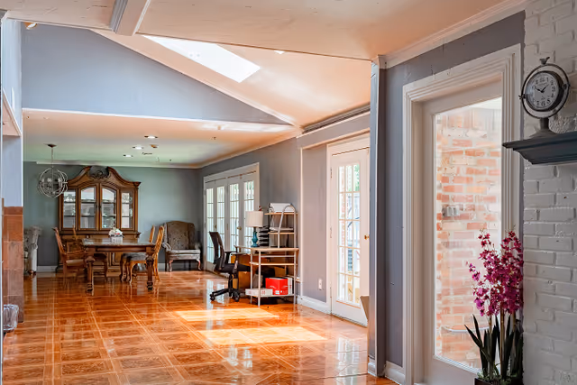 Bright open living and dining area with glossy hardwood floors, a dining table and china cabinet at the far end, a desk by glass doors, and a potted orchid by a white brick fireplace.