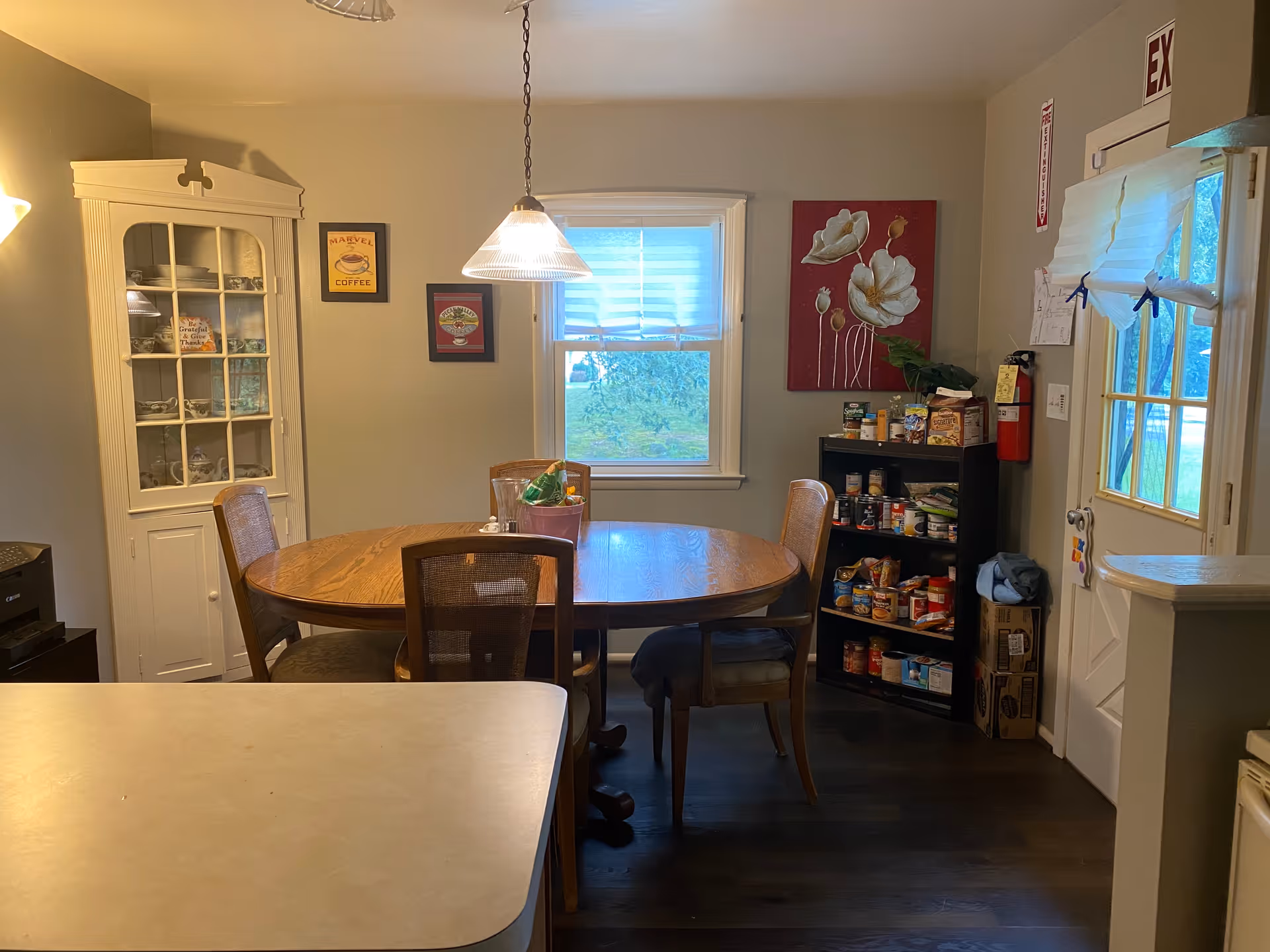 A dining room with a wooden oval table surrounded by four chairs. A hanging light fixture is above the table. To the left, there is a white corner cabinet with glass doors displaying dishes. On the right side, there is a black shelving unit stocked with canned and boxed food items next to a door with a window and an exit sign above it. The walls are decorated with framed pictures and a large floral painting. A window with blinds is centered on the back wall.