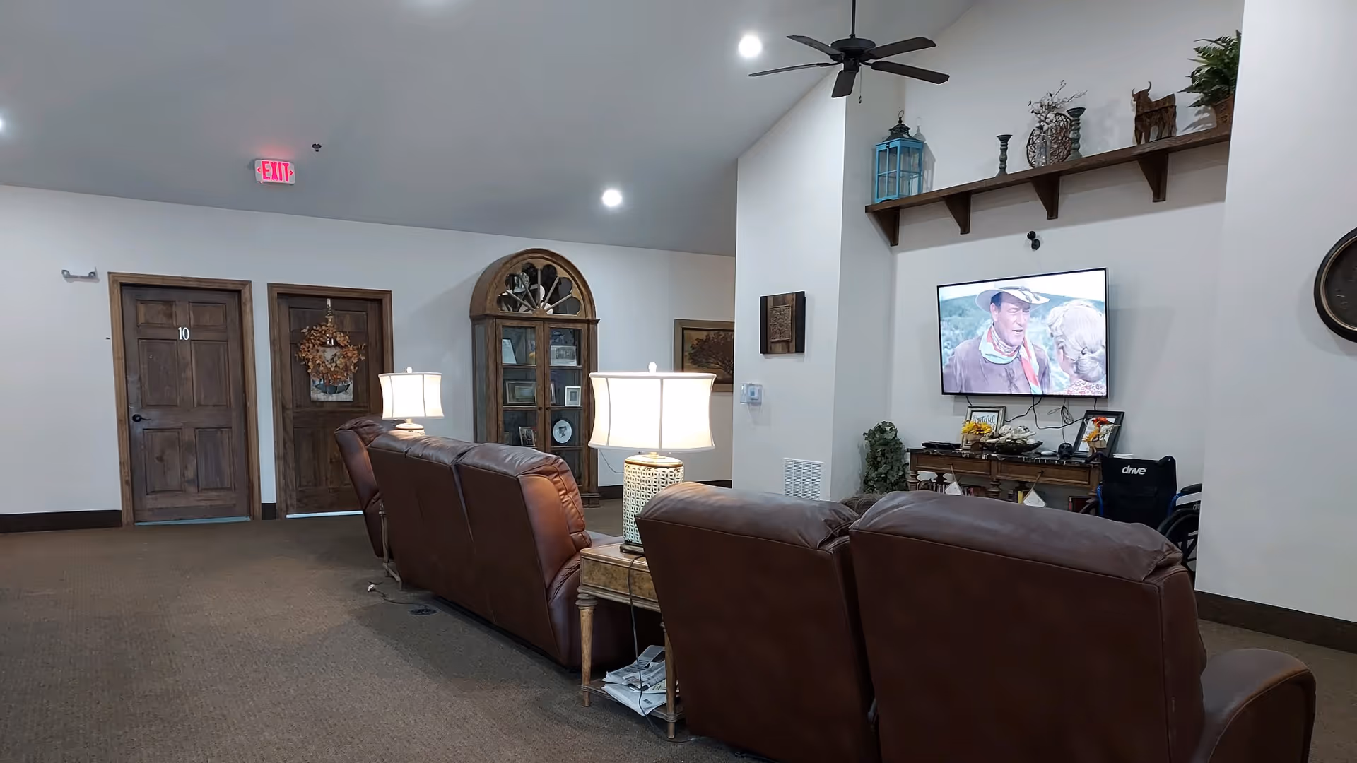 A cozy living room area in a senior living facility with three brown leather recliners facing a wall-mounted TV showing a western movie. There are two table lamps on side tables between the recliners. The room has a high ceiling with recessed lighting and a ceiling fan. On the wall behind the TV, there is a wooden shelf with decorative items and plants. To the left, there are two wooden doors, one marked with the number 10 and the other decorated with a wreath. A wooden cabinet with glass doors and framed pictures is also visible.