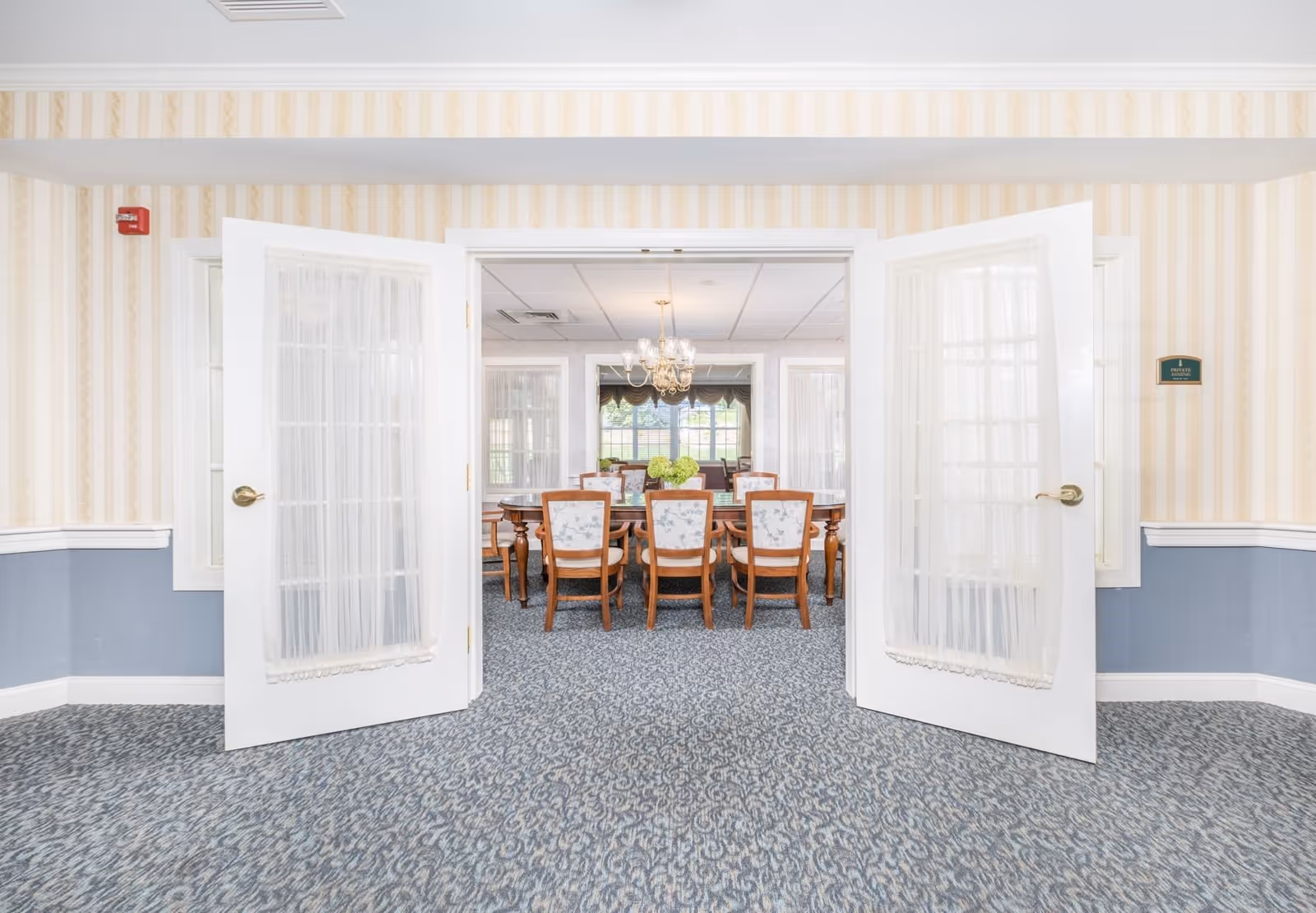 View through open double doors into a private dining room with a wooden dining table and eight chairs with floral upholstery. The room has patterned carpet, wallpaper with vertical stripes, a chandelier, and large windows with curtains.