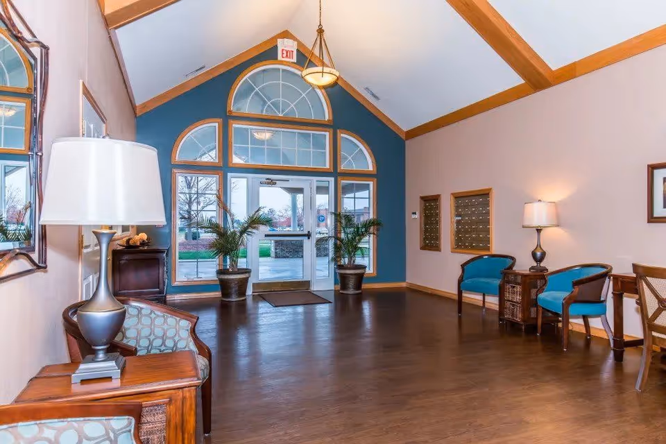 Interior view of a senior living facility lobby with wooden floors, a high vaulted ceiling with wooden beams, large arched windows and glass door entrance. The room is furnished with several chairs, side tables with lamps, two potted plants near the entrance, and a mirror on the left wall.