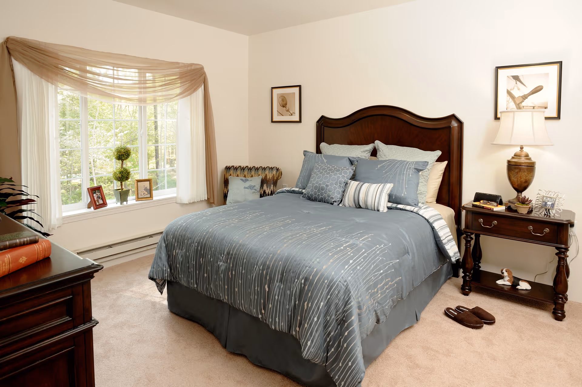 A cozy bedroom with a large wooden bed featuring blue and gray bedding and multiple pillows. There is a wooden nightstand with a lamp, books, and decorative items next to the bed. A pair of slippers is placed on the carpeted floor beside the nightstand. A large window with sheer white curtains and a light brown swag valance lets in natural light. The room also has framed pictures on the walls and a wooden dresser partially visible on the left side.