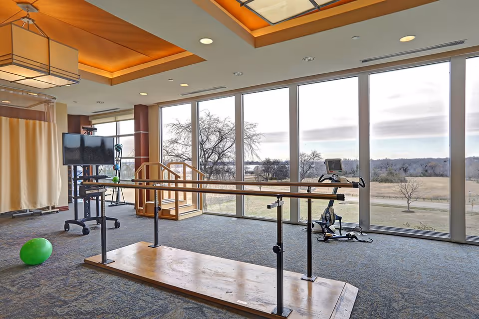 A spacious rehabilitation or physical therapy room with large floor-to-ceiling windows showing an outdoor view. The room contains parallel bars for walking exercises, a stationary exercise bike, a green exercise ball on the carpeted floor, a TV on a mobile stand, and a small set of wooden stairs with handrails. The ceiling has recessed lighting and a decorative light fixture.