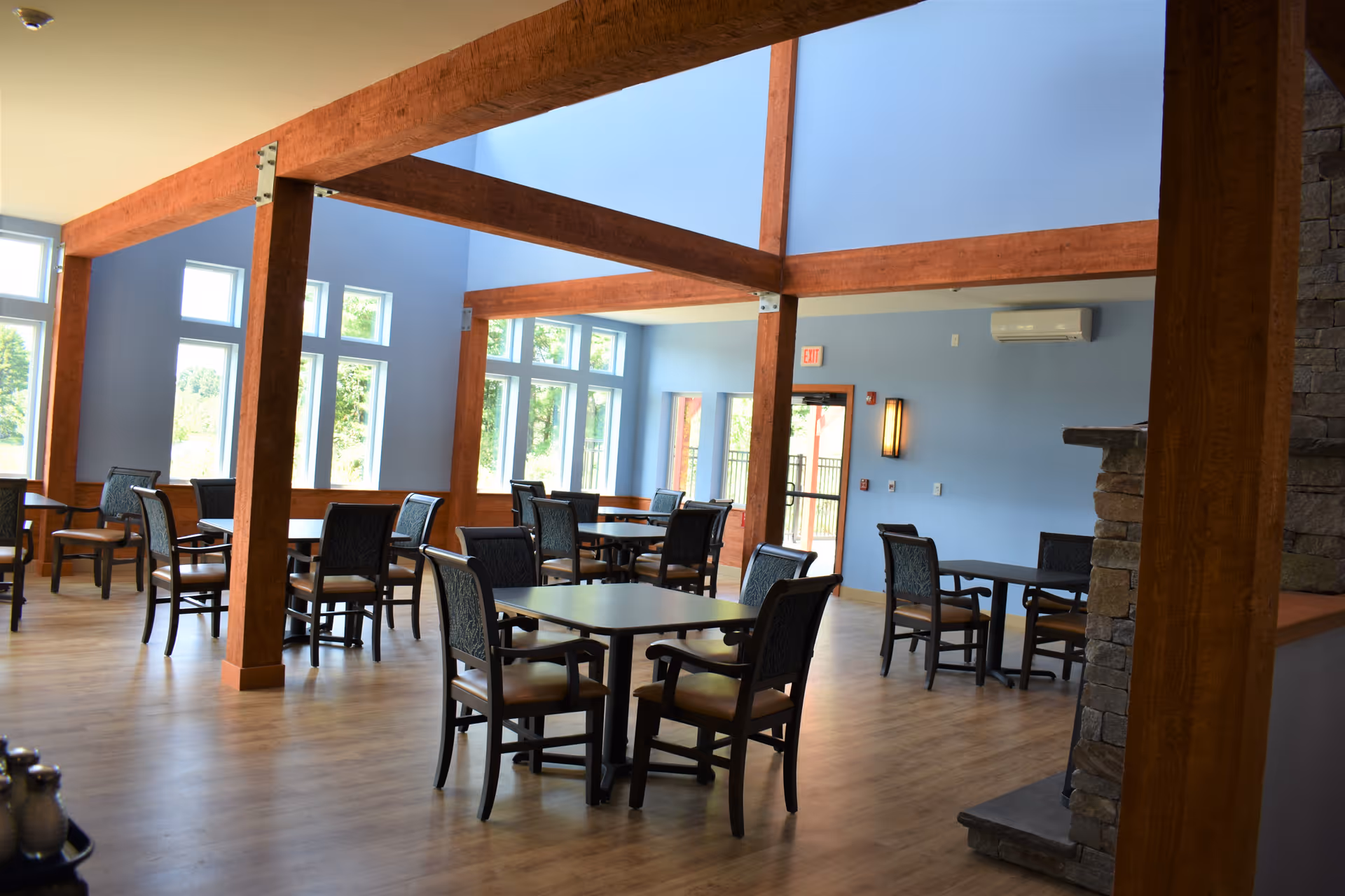 A spacious dining area with multiple tables and chairs arranged neatly. The room features large windows allowing natural light to fill the space, wooden beams on the ceiling, light blue walls, and a stone fireplace on the right side.