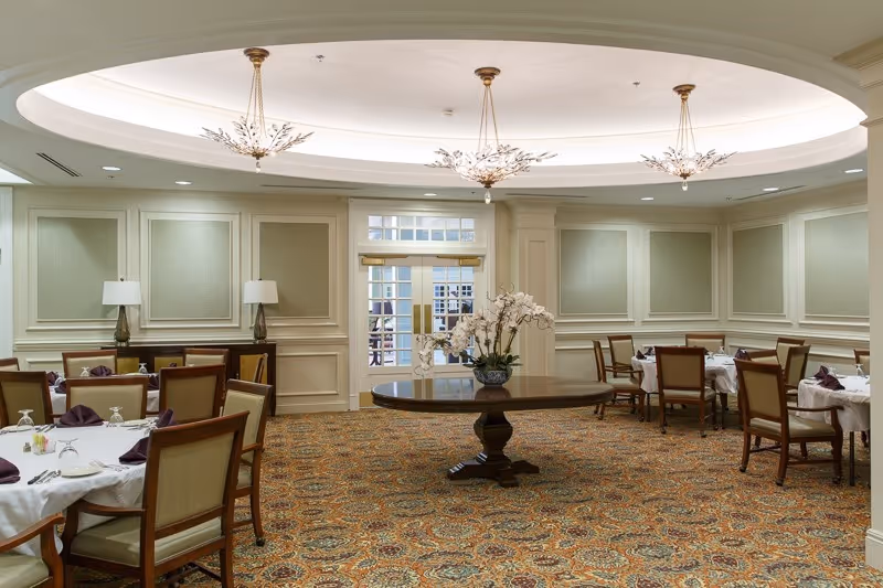 A well-lit dining room with round tables covered in white tablecloths, each set with folded purple napkins, glasses, and silverware. The room features a patterned carpet, beige walls with decorative paneling, two table lamps on a sideboard, and three elegant chandeliers hanging from a recessed ceiling. A round wooden table with a floral arrangement is centered in the room, and double glass doors are visible at the far end.
