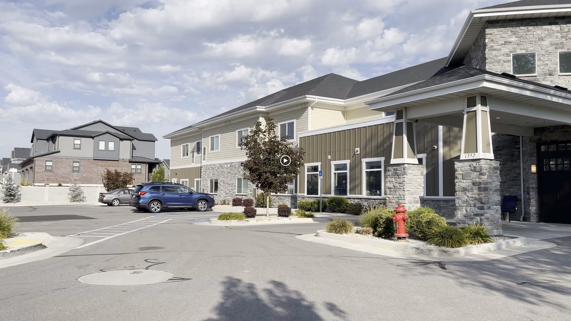 Front entrance and parking area of a two-story assisted living facility with stone pillars, landscaping, and parked cars under a partly cloudy sky.