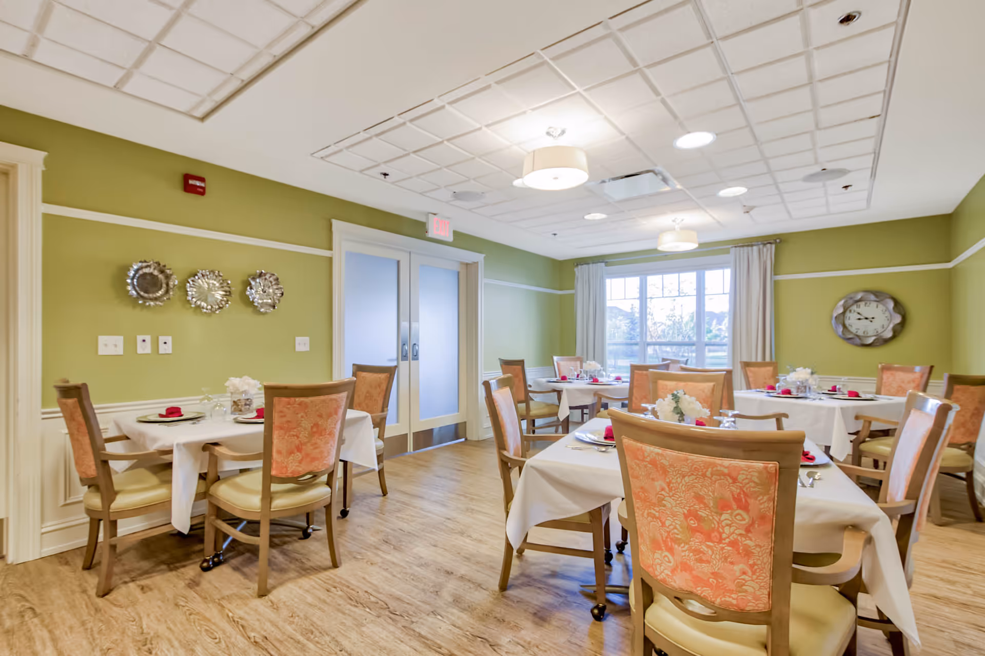 A dining room in a senior living facility with several tables covered in white tablecloths, each set with plates, glasses, and red napkins. The room has green walls, wooden flooring, and large windows with white curtains allowing natural light to enter. There are decorative plates on one wall and a clock on another. The ceiling has recessed lighting and hanging light fixtures.