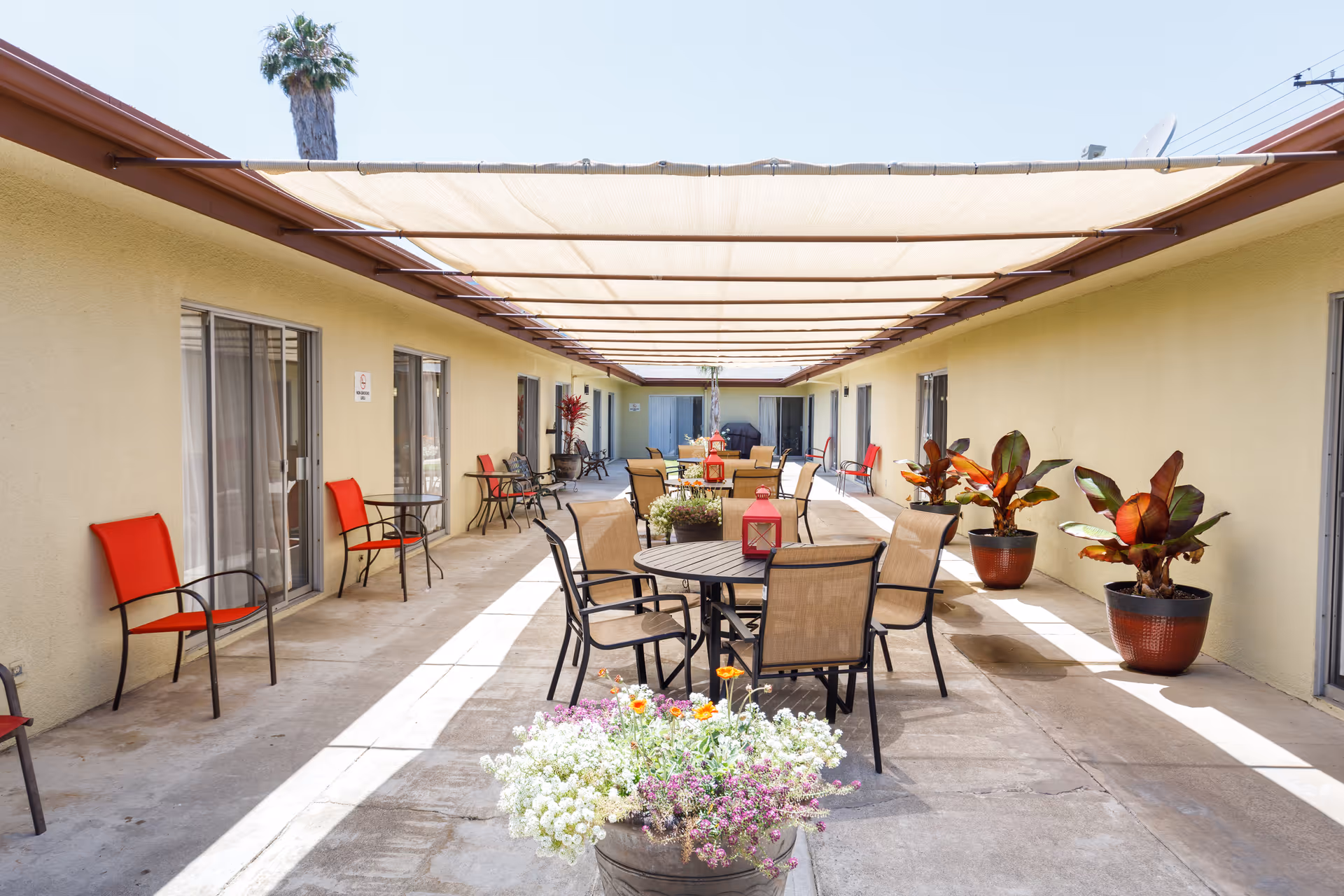 Outdoor covered patio area at Mission Palms Healthcare Center with multiple tables and chairs arranged for seating. The space is shaded by a fabric canopy overhead, with potted plants and flowers adding greenery along the sides. Doors to rooms or units line both sides of the walkway.