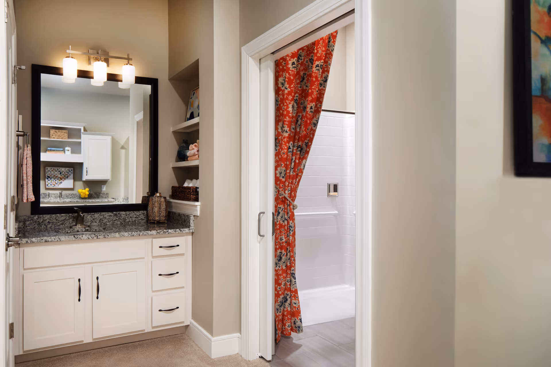A bathroom vanity area with a granite countertop, white cabinets, a large mirror with three light fixtures above it, and open shelves with towels and decorative items. To the right, there is a doorway with a red floral shower curtain partially open, revealing a white tiled shower area.