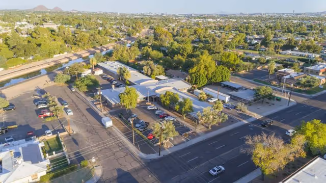 Aerial view of Desert Terrace Healthcare Center showing the building complex surrounded by trees, parking lots with several cars, and adjacent streets. The surrounding area includes residential neighborhoods and greenery extending into the distance.
