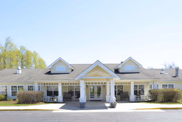 Front exterior view of a single-story building with a covered entrance supported by white columns, surrounded by bushes and trees under a clear blue sky.