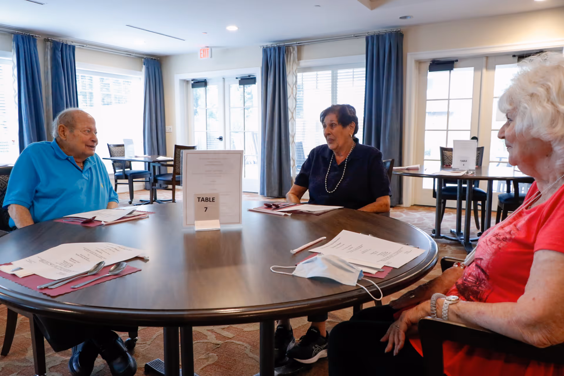 Three elderly people sitting around a round dining table in a well-lit room with large windows and curtains. The table has menus, utensils, and a sign that reads 'TABLE 7'. One person is wearing a blue shirt, another a dark top with a necklace, and the third a red shirt. A face mask is placed on the table.