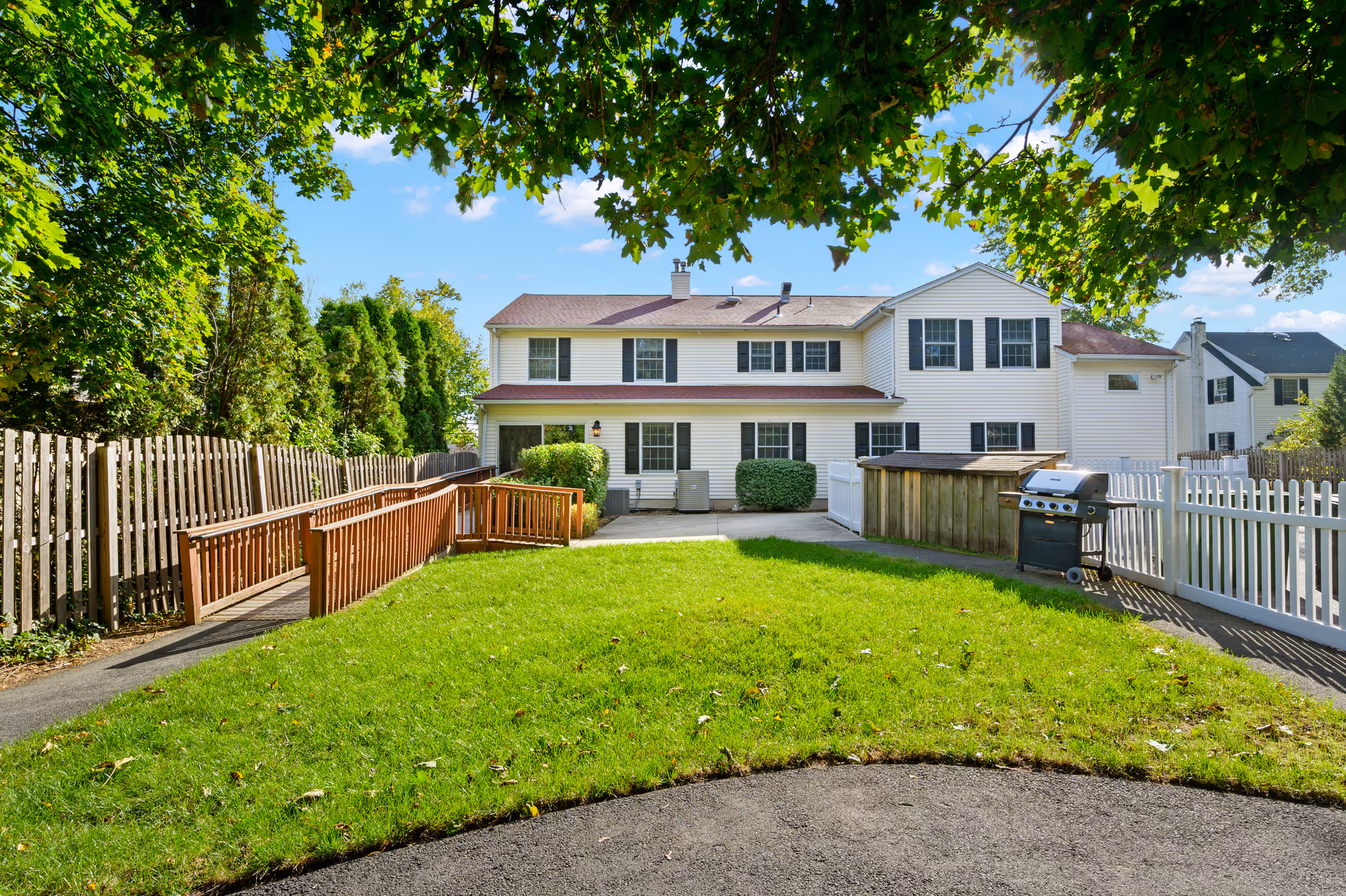 Rear exterior of a two-story white building with a grassy yard, wooden accessibility ramp, and a barbecue grill under leafy trees.
