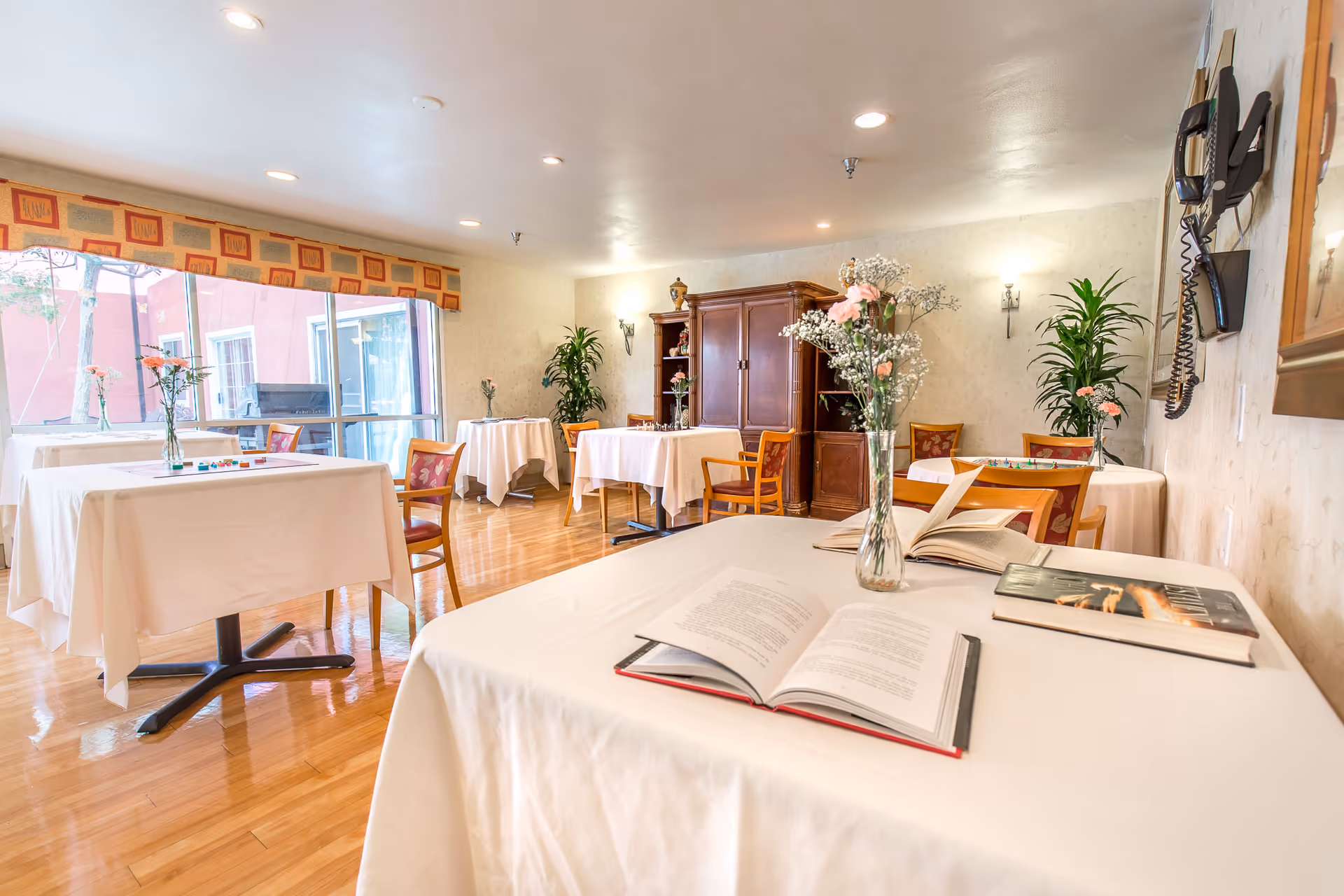 A bright dining room with several tables covered in white tablecloths, each adorned with a vase of flowers. There are wooden chairs around the tables, a large window letting in natural light, and a wooden cabinet against the far wall. An open book and a closed book are on the table in the foreground.