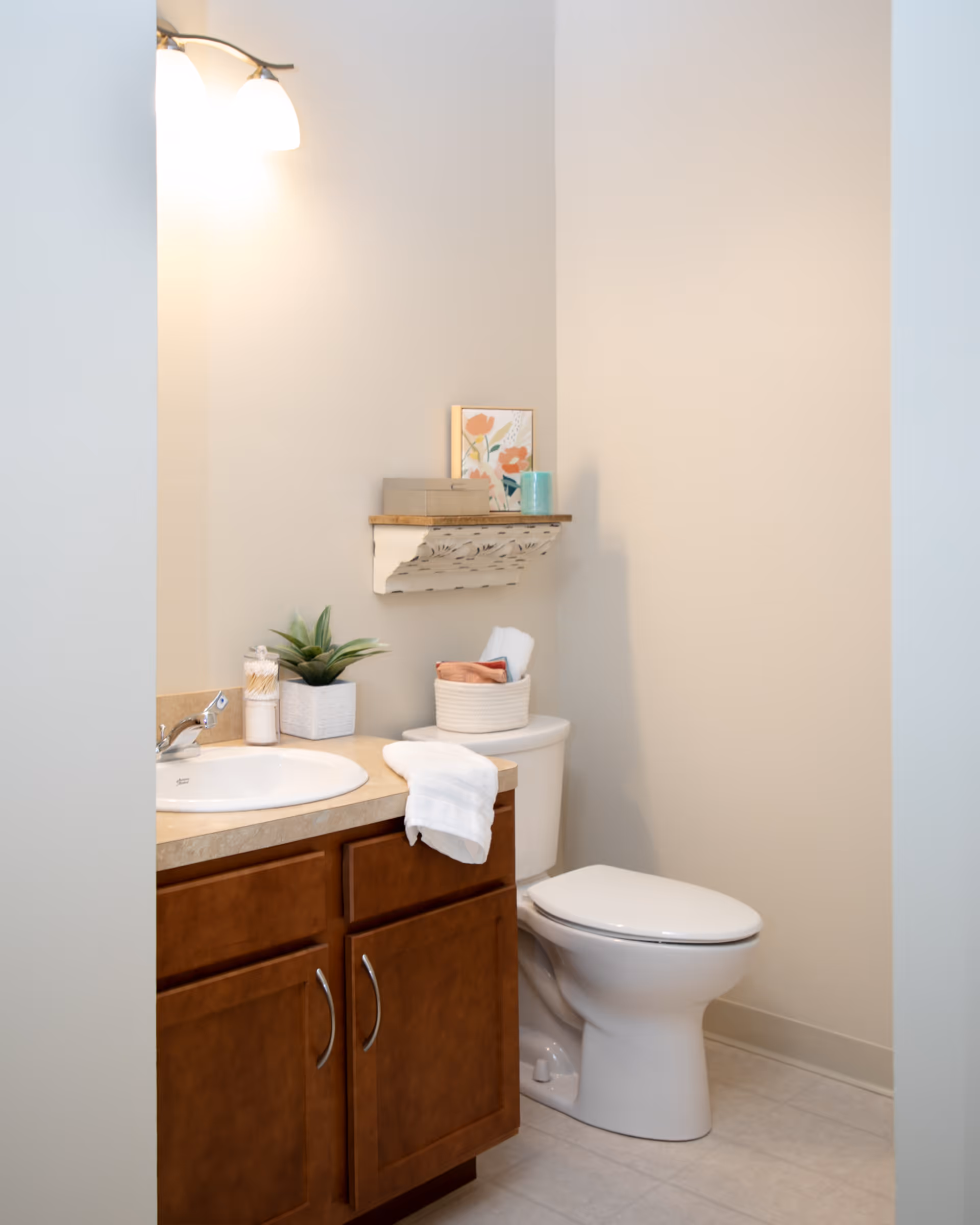 A clean bathroom with a wooden vanity cabinet, a white sink, and a toilet. Above the toilet is a decorative shelf holding a framed floral picture, a small box, and a teal cup. A white towel hangs over the edge of the vanity, and a small potted plant and cotton swabs are on the countertop. The walls are painted a light beige color.