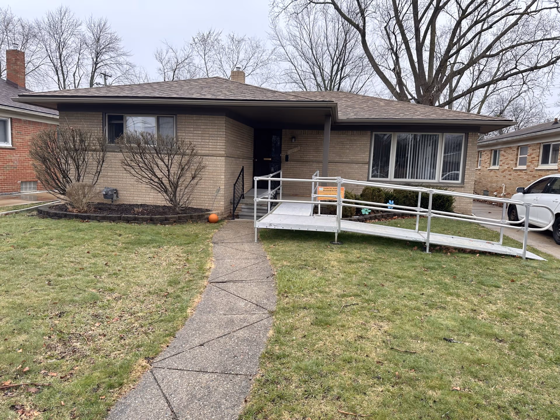 Front exterior view of a single-story brick residential building with a sloped roof, a concrete pathway leading to the entrance, and a metal wheelchair ramp on the right side. There are leafless trees and shrubs around the building, and a white car is parked on the driveway to the right.