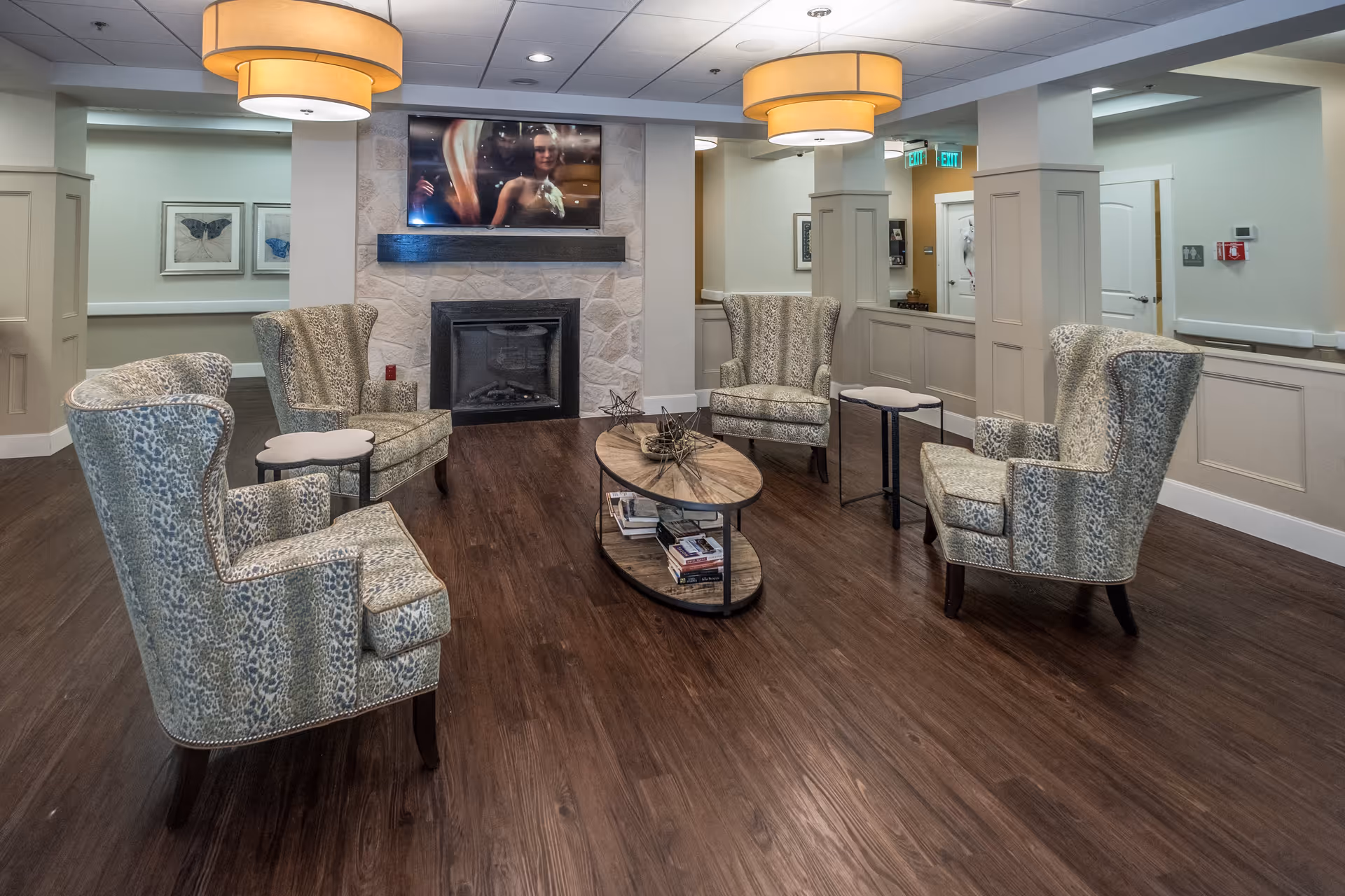 A cozy sitting area in a senior living facility with four patterned armchairs arranged around an oval wooden coffee table. The table has decorative items and books on its lower shelf. A stone fireplace with a mounted flat-screen TV above it is visible on the far wall. The room has warm lighting from two large ceiling lamps and features wood flooring and neutral-colored walls with wainscoting.