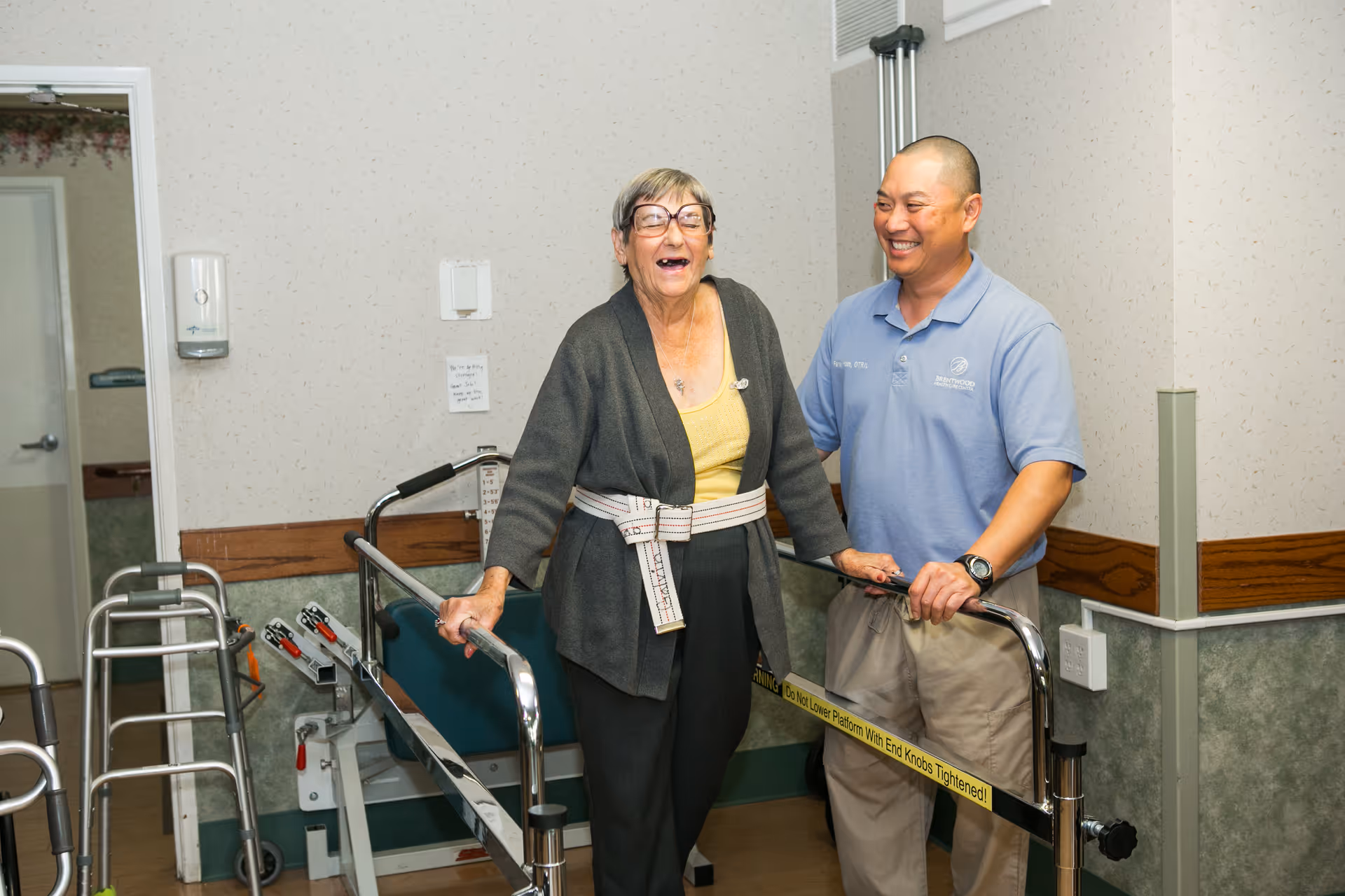 An elderly woman wearing glasses and a support belt is using parallel bars for walking assistance while a smiling male caregiver in a blue polo shirt stands beside her, providing support in a healthcare facility hallway.