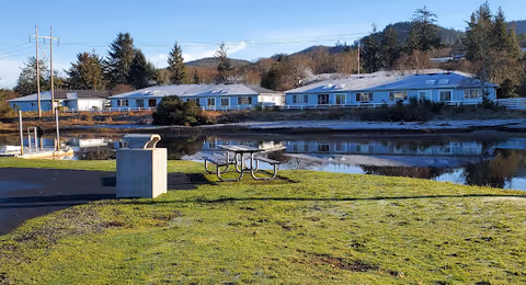 Outdoor view of a senior living facility named Neawanna By The Sea, showing a grassy area with a picnic table and a trash bin near a calm body of water, with single-story buildings and trees in the background under a clear sky.
