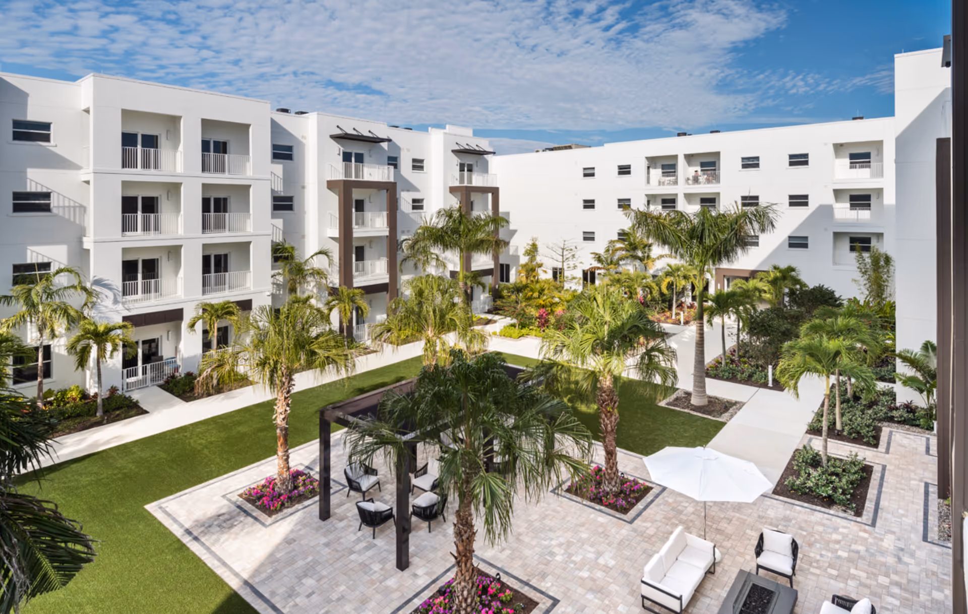 Outdoor courtyard area of a senior living facility with white multi-story buildings surrounding a landscaped garden. The courtyard features palm trees, green grass, paved walkways, and seating areas with chairs and a white umbrella under a pergola.