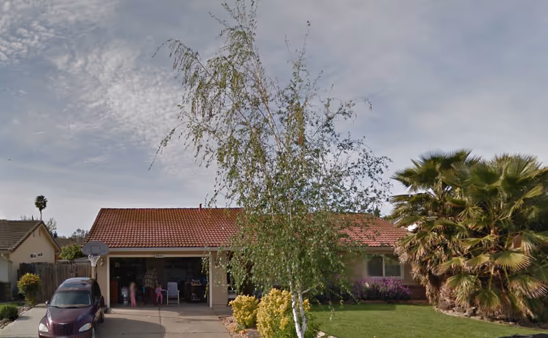 Front exterior view of a single-story house with a red tiled roof, a driveway with a parked car, a basketball hoop, a tree in the front yard, and palm trees on the right side under a partly cloudy sky.