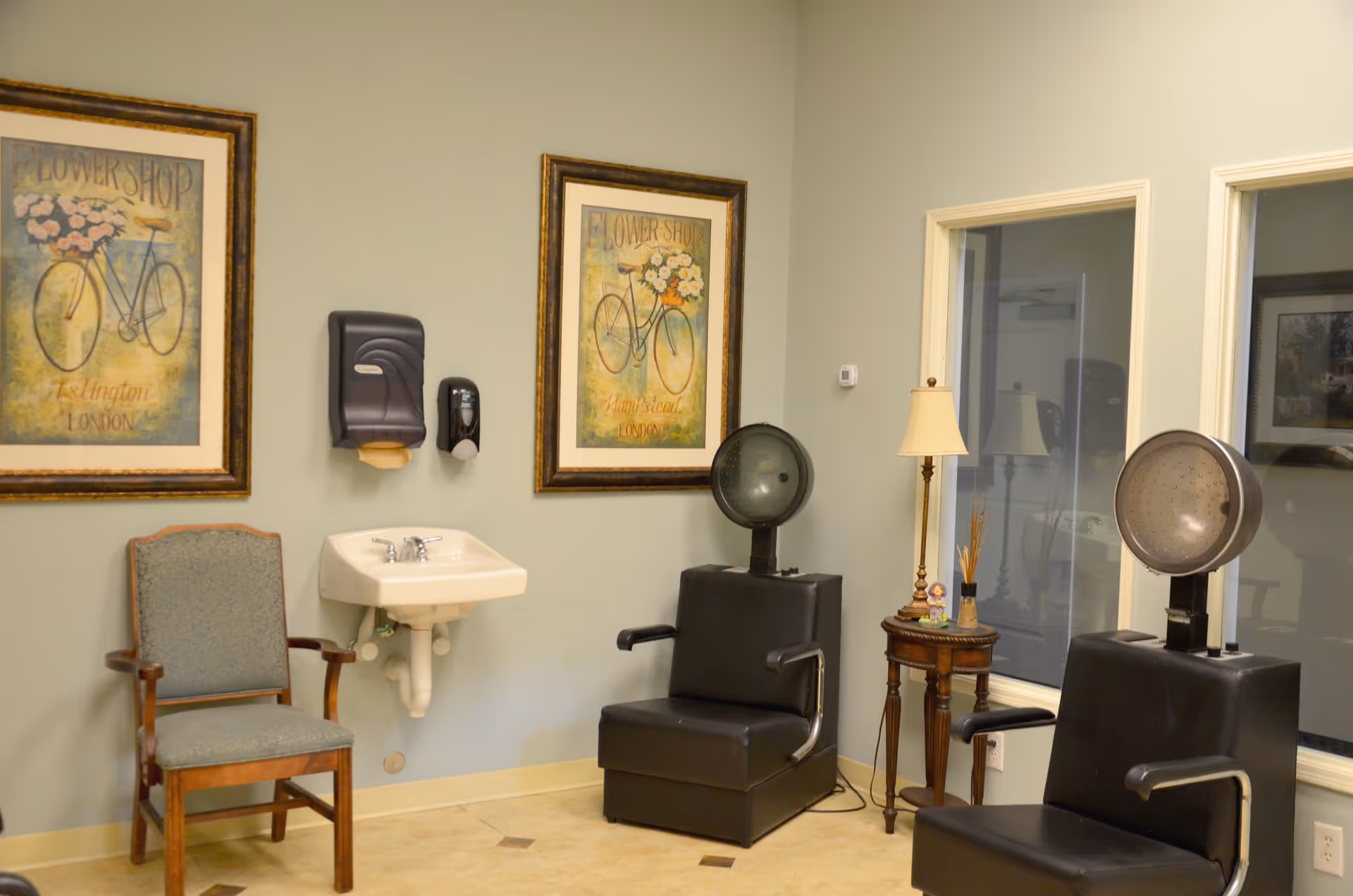 Small salon room with two black hooded hair-dryer chairs, a wall-mounted sink, framed bicycle prints, and a waiting chair.