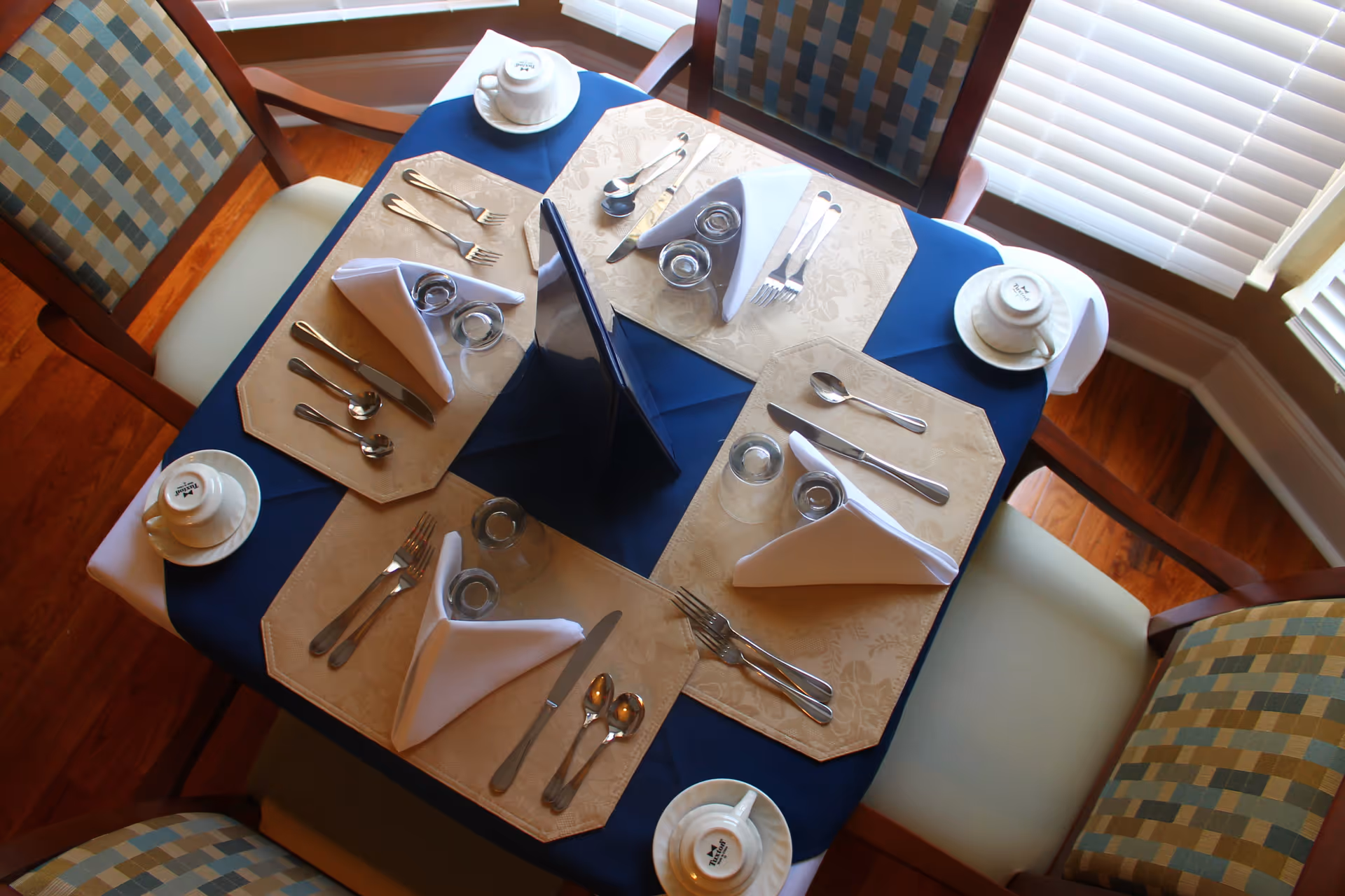 A dining table set for four with beige placemats, folded white napkins, silverware, glasses, and white cups and saucers. The table has a dark blue tablecloth and is surrounded by four cushioned chairs with patterned upholstery. The setting is near windows with white blinds.