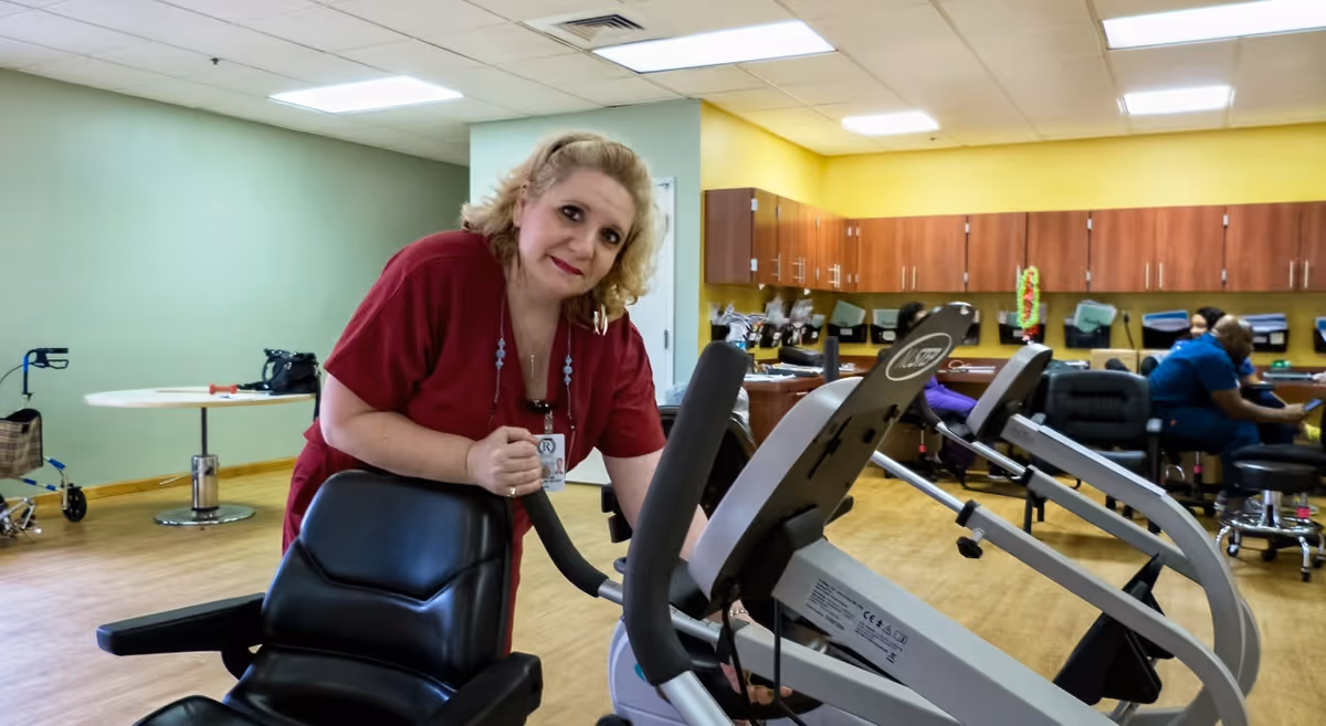 A staff member in a red uniform leans on exercise equipment in a communal activity/therapy room with cabinets and other people in the background.