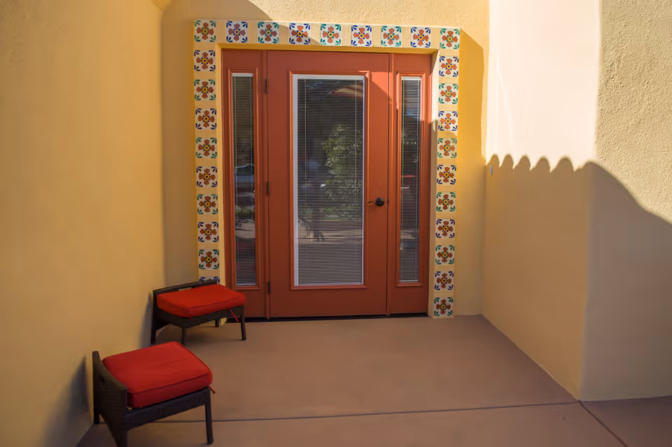 A small outdoor patio area with two wicker chairs featuring red cushions, positioned near a set of glass double doors framed with decorative colorful tiles. The walls and floor are painted in warm yellow and beige tones, with sunlight casting shadows on the right wall.