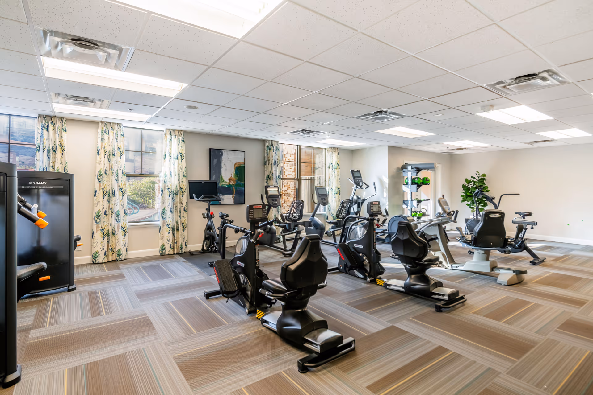 A bright and spacious fitness room with several exercise bikes, elliptical machines, and weight equipment. The room has large windows with patterned curtains allowing natural light to enter. The floor is covered with patterned carpet tiles, and there is a rack with green dumbbells and a potted plant in the corner.