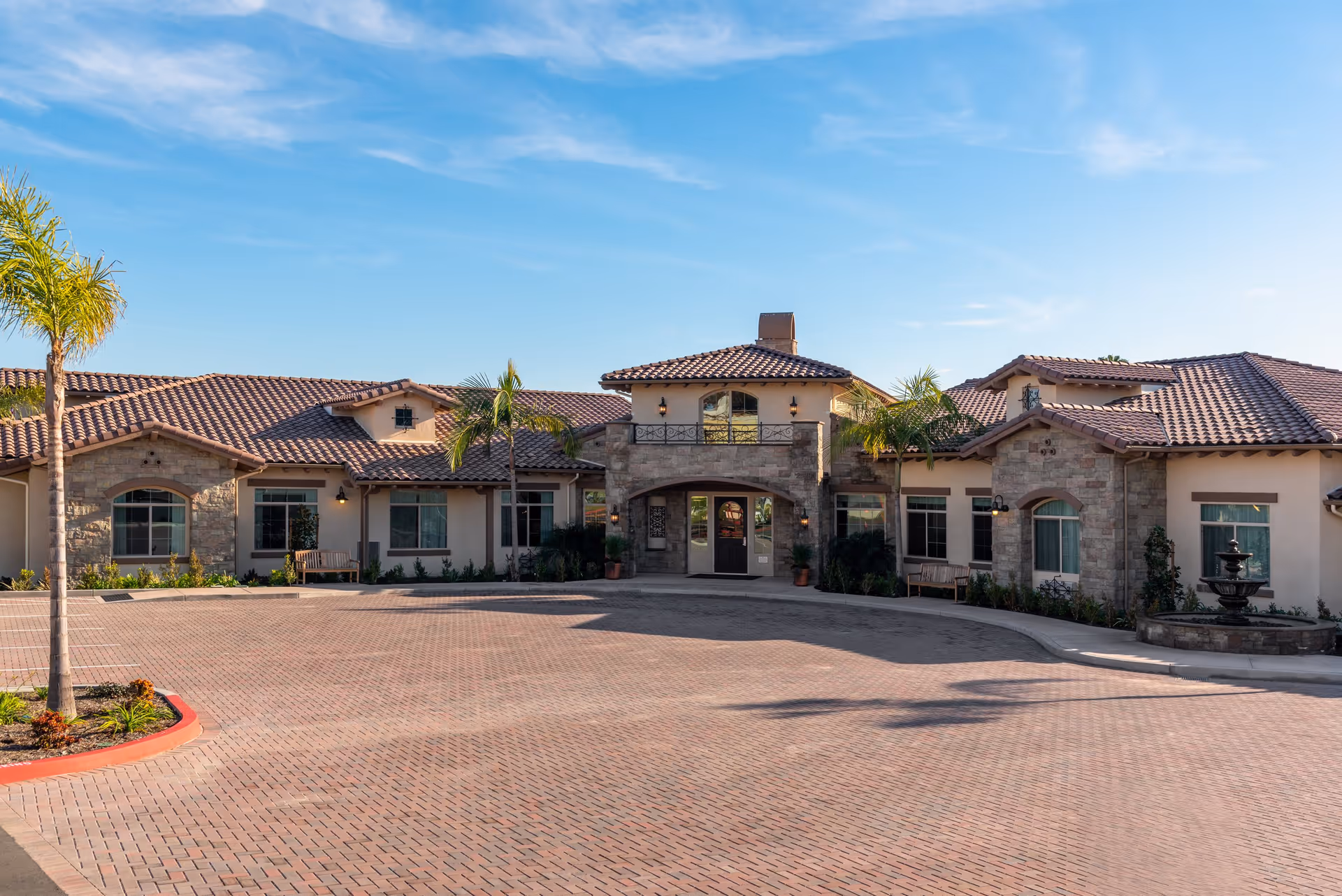 Front exterior view of ActivCare Laguna Hills facility featuring a single-story building with a tiled roof, stone and stucco facade, palm trees, benches, and a fountain under a clear blue sky.