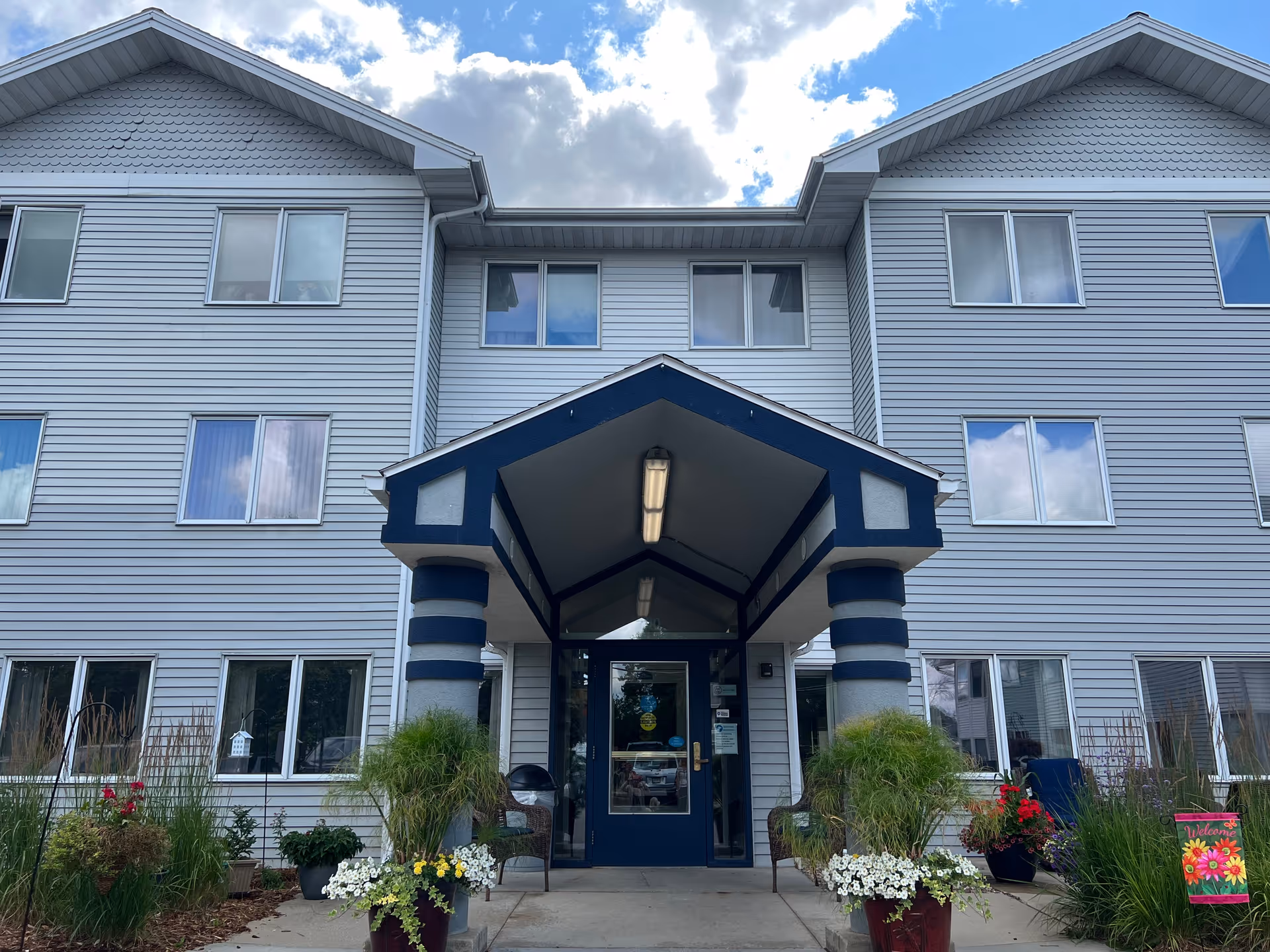 Front exterior view of a three-story building with gray siding and multiple windows. The entrance has a blue and white covered porch supported by two columns. There are potted plants and flowers on either side of the entrance walkway, and a small colorful welcome sign is visible on the right side near the plants. The sky is partly cloudy.