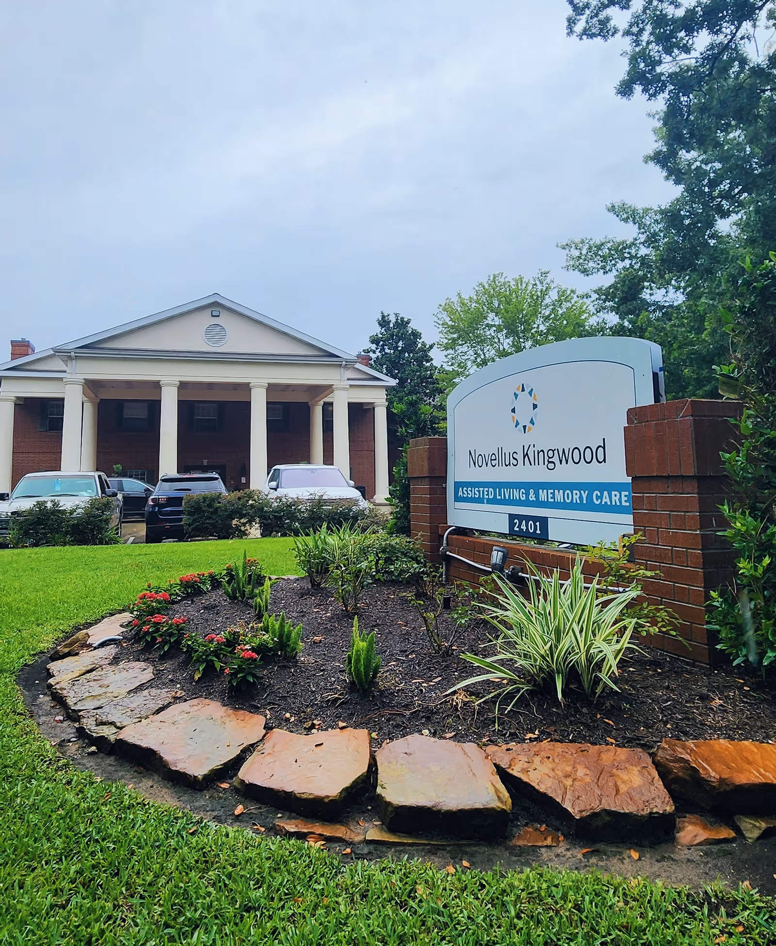 Front exterior of Novellus Kingwood assisted living facility with a large entrance, landscaped flowerbed, parked cars, and a prominent sign.