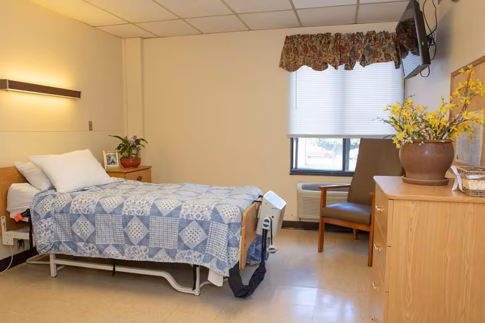A simple, clean bedroom in a senior living facility with a single bed covered by a blue and white patterned quilt. There is a wooden nightstand with a potted plant and a framed photo next to the bed. A wooden dresser with a large potted yellow flower arrangement sits near a window with blinds and a floral valance. A brown chair is positioned near the window, and a flat-screen TV is mounted on the wall above the dresser.