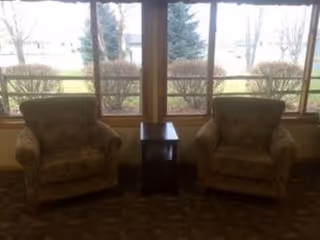 Two upholstered armchairs flanking a small wooden side table in front of large windows overlooking a lawn and trees.