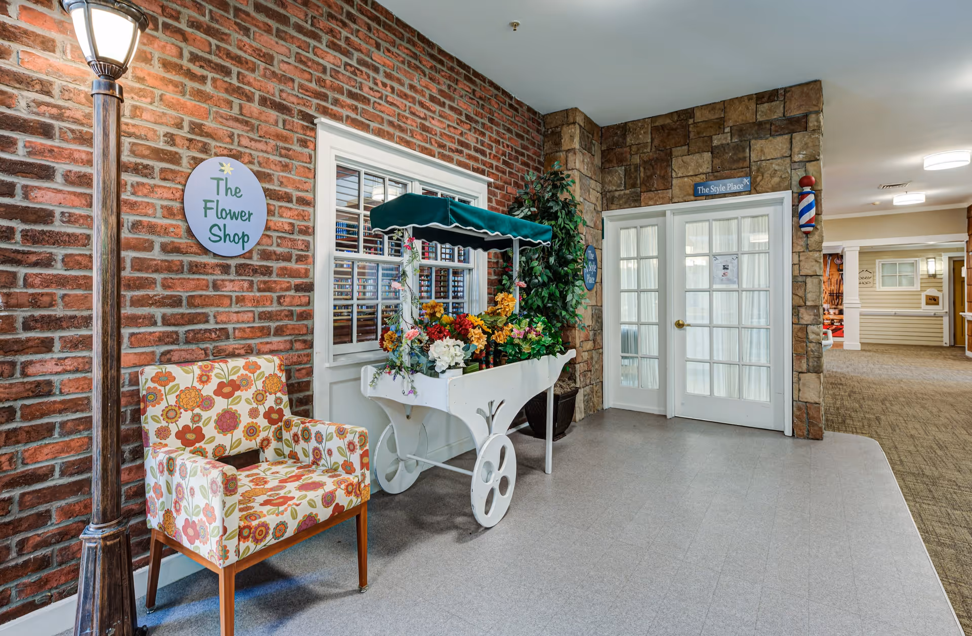 Indoor hallway area with a brick wall on the left featuring a floral patterned armchair and a white flower cart filled with colorful artificial flowers under a green canopy. A sign on the brick wall reads 'The Flower Shop'. To the right, there is a stone wall with double glass doors labeled 'The Style Place' and a traditional barber pole mounted on the wall. The hallway extends further with carpeted flooring and additional doors and decor visible in the background.